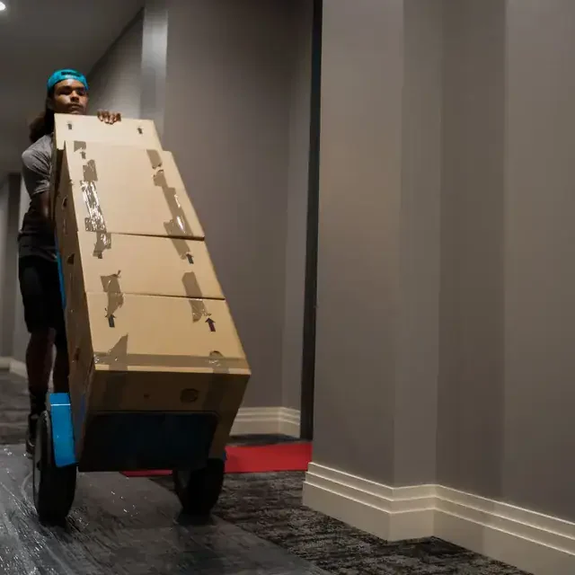 Man-moving-large-box-on-dolly-in-hallway Professional mover from BEK Moving Co. using a dolly to transport stacked moving boxes through a residential hallway.